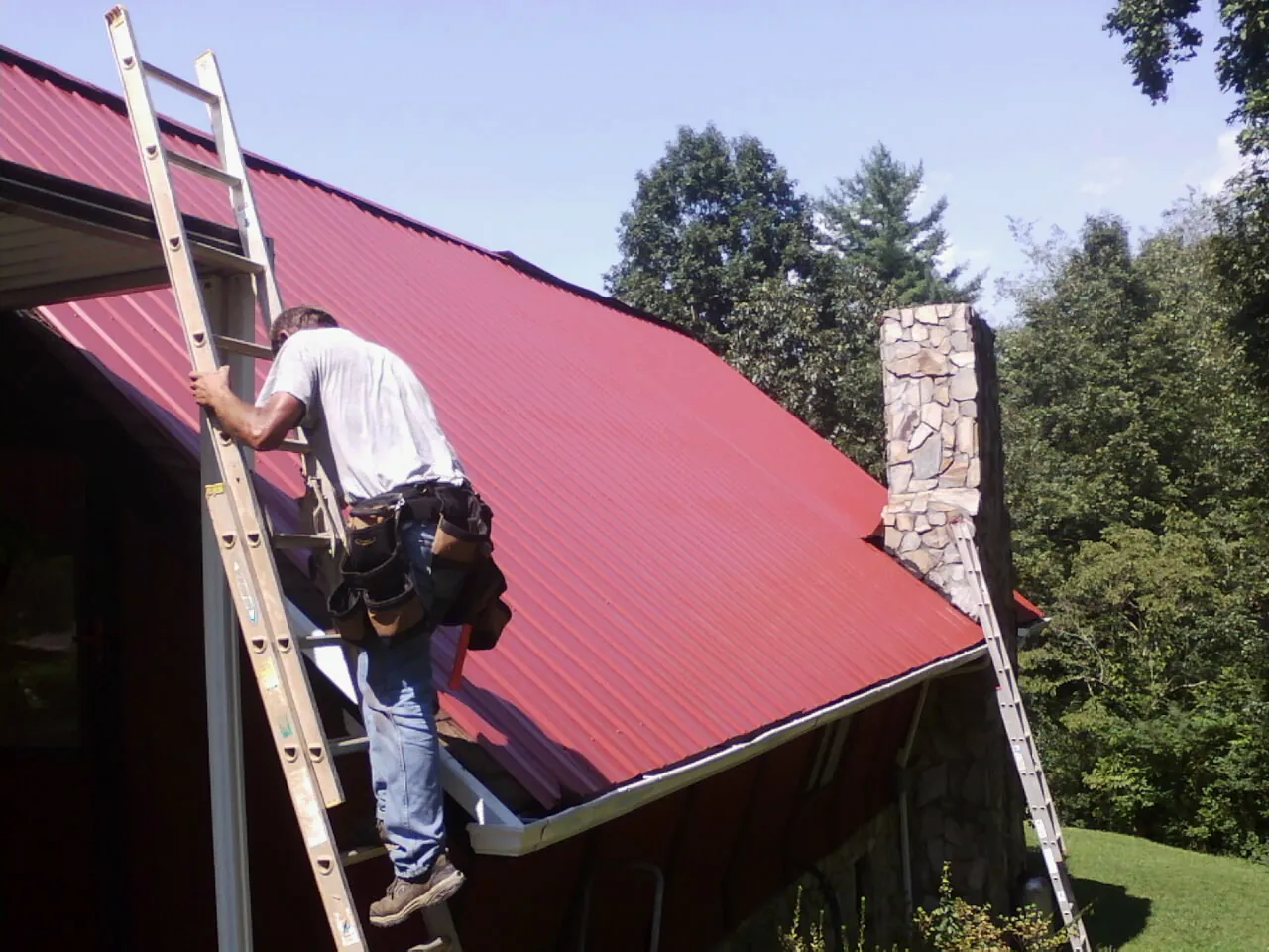 Roofer on a ladder beside a red metal roof and stone chimney — Soffit & Fascia Repair by Roofing Brothers in Athens